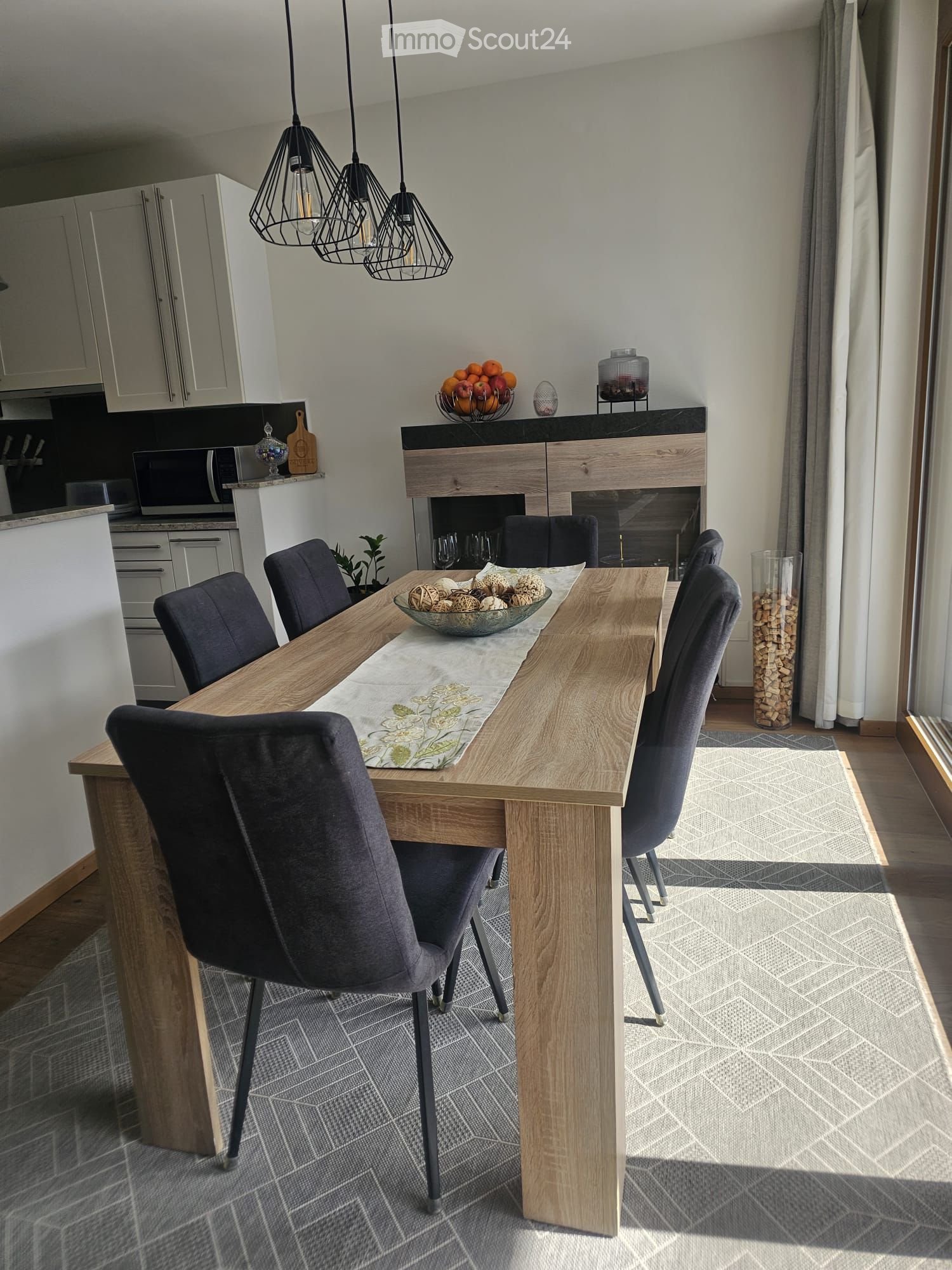 Modern dining area with a wooden dining table, six dark grey chairs, a decorative bowl with decorative items, a fireplace, and pendant lights. The area is connected to a kitchen with white cabinets and a microwave.