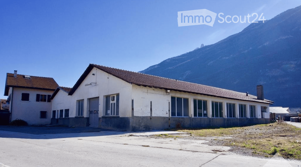 A two-story building with a flat roof and large windows, situated in a rural area with a mountain in the background.