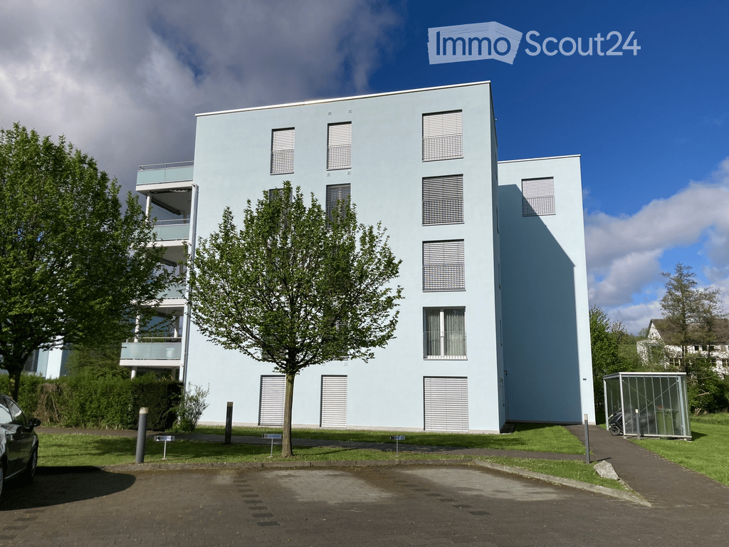 Modern, light blue apartment building with multiple floors, balconies, and large windows. The building is surrounded by greenery and has a paved parking area.
