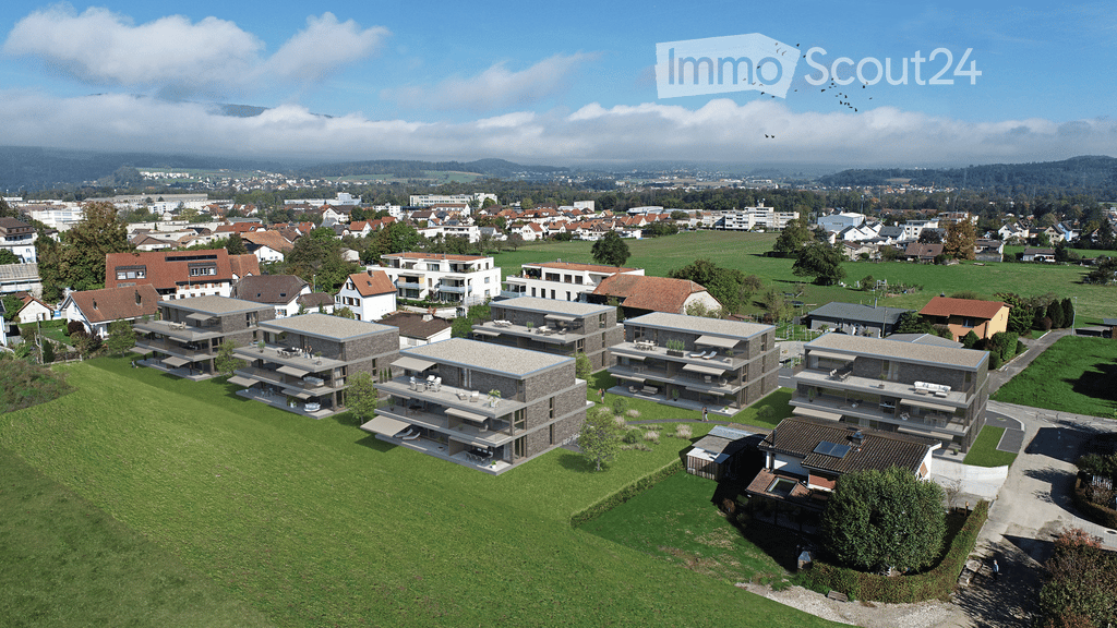 Aerial view of a residential area with modern apartment buildings, green lawns, and surrounding town with houses and fields.
