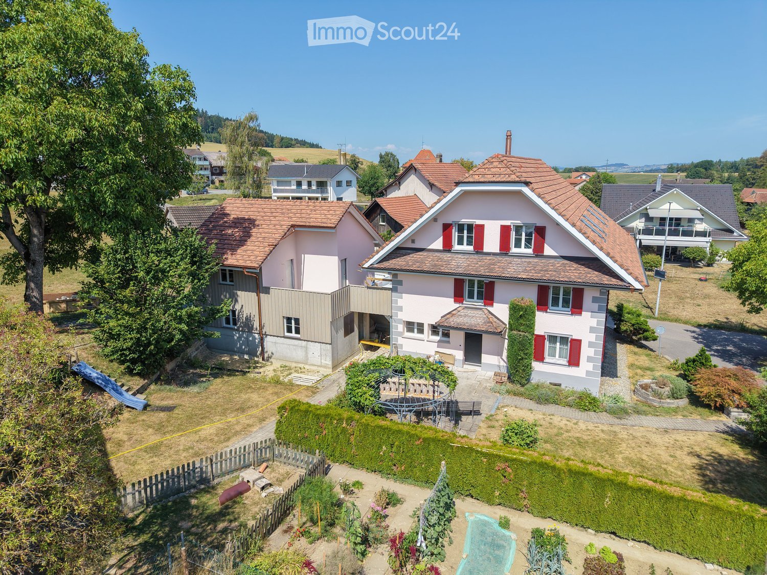 A two-story house with a red-tiled roof and white walls, surrounded by a well-maintained garden and other houses in the background.