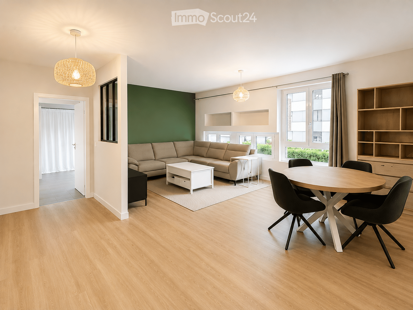 Modern living and dining area with light wood flooring, a beige sectional sofa, a white coffee table, a round wooden dining table with four black chairs, and built-in wooden shelving units.
