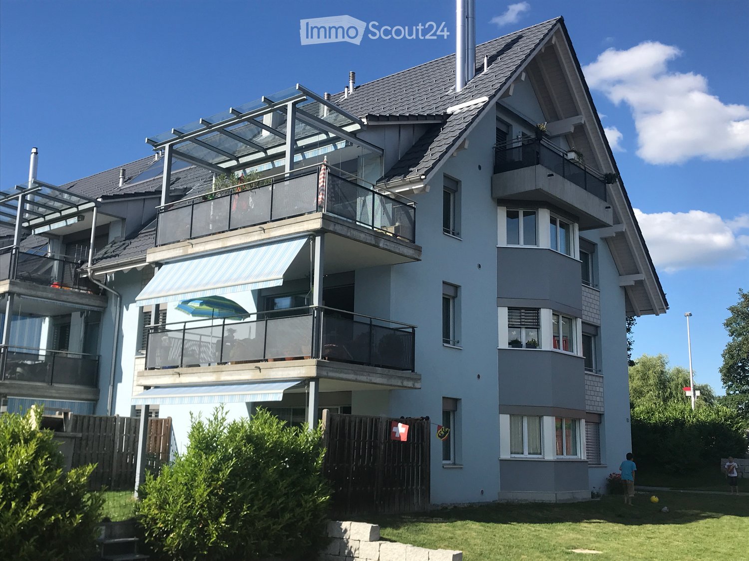 Two-story house with multiple balconies, large windows, and a covered terrace area. The house features a modern design with grey and white exterior walls, multiple chimneys, and a well-maintained garden in front.