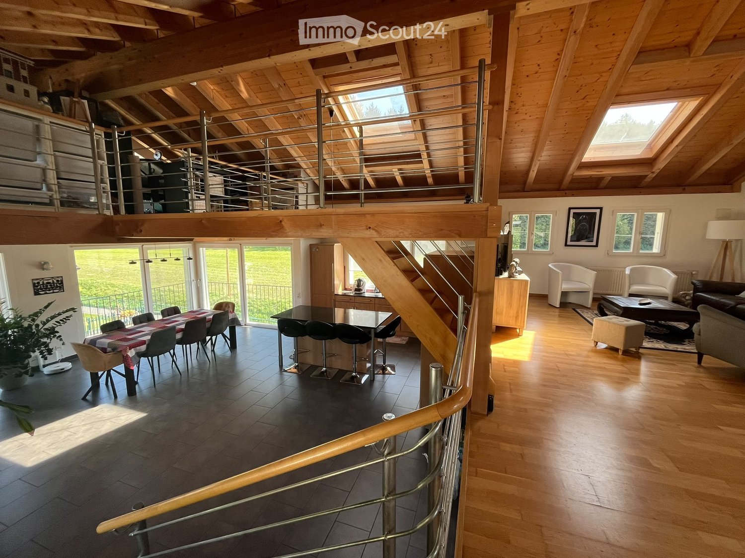 Open-plan living and dining area with wooden beams, a staircase leading to an upper level, and large windows offering a view of the greenery outside. The living area features a sofa, chairs, a coffee table, and a floor lamp. The dining area includes a din