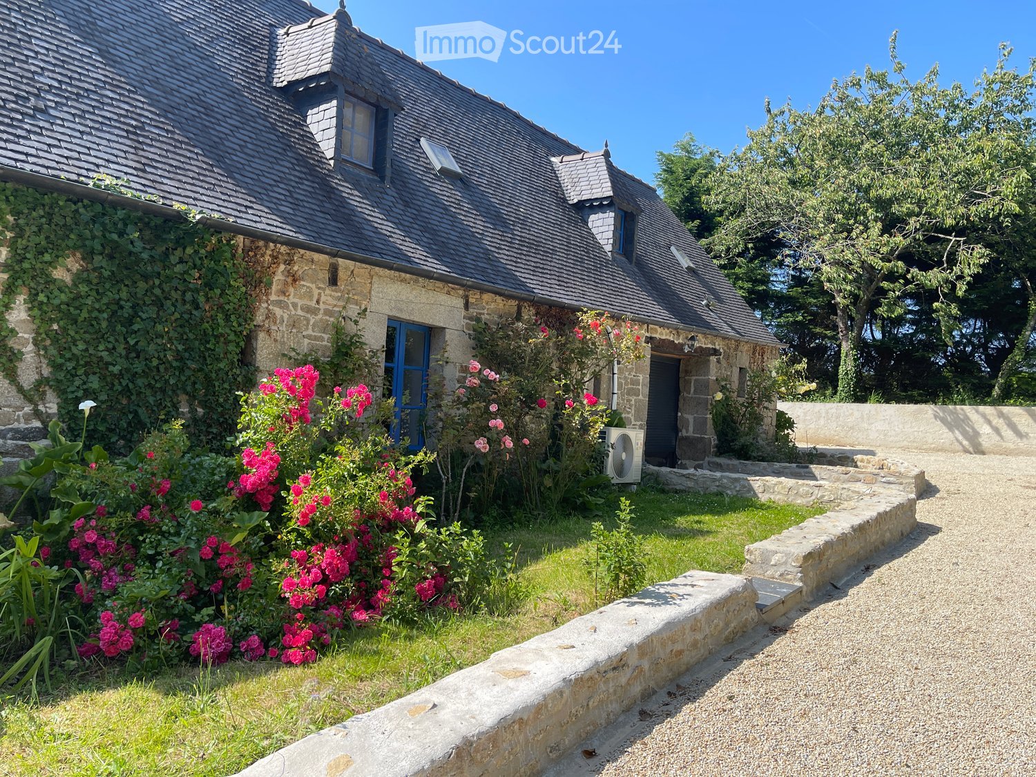 Stone house with a dark slate roof, surrounded by a garden with pink flowers and a stone pathway leading to the entrance.