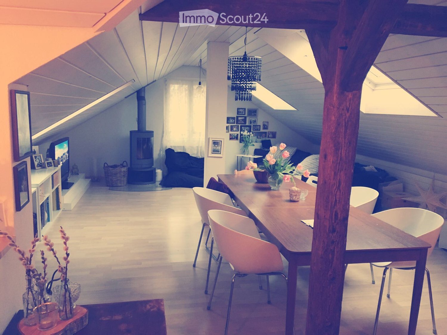 Bright, open loft-style room with exposed wooden beams, skylights, and a fireplace. Features a long wooden dining table with modern chairs, a sofa, a TV, and decorative vases with flowers.