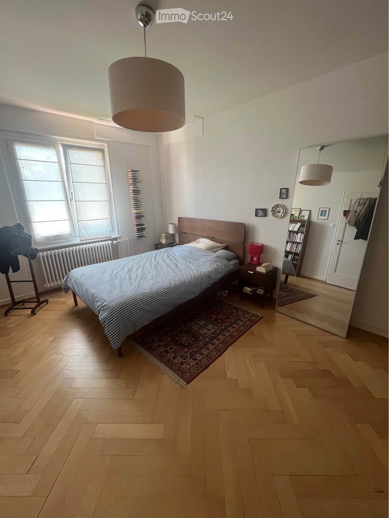 Well-lit bedroom with wooden flooring, large window, bed with striped bedding, nightstand, bookshelf, and mirror.