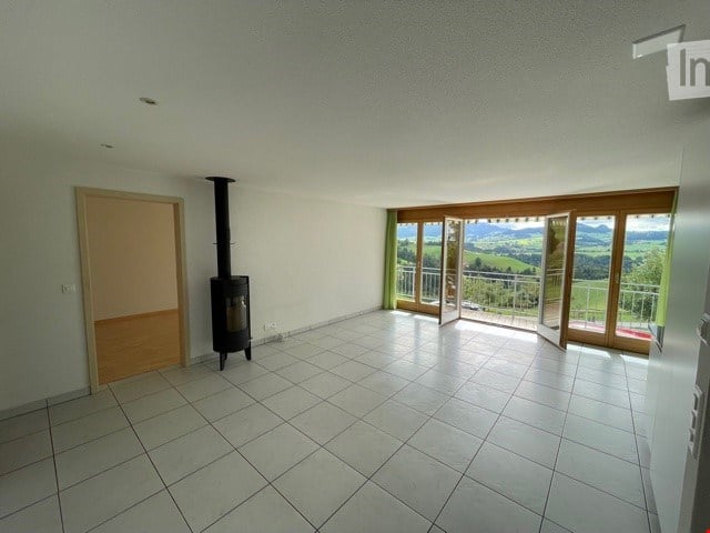 Empty room with tiled floor, white walls, a wood-burning stove, and large windows offering a scenic view of the landscape.