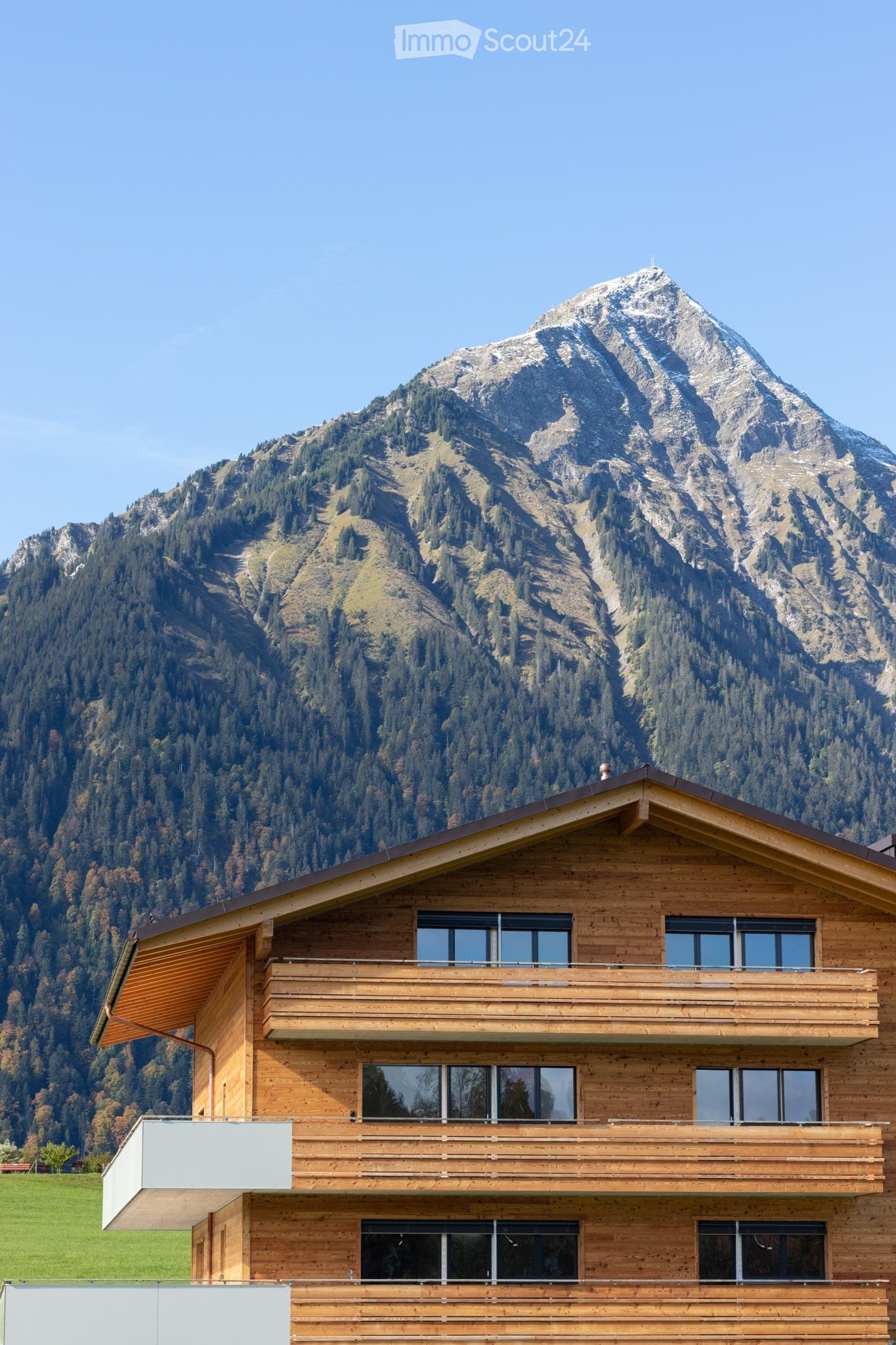 A modern wooden house with multiple balconies and large windows, set against a backdrop of a snow-capped mountain and forested landscape.