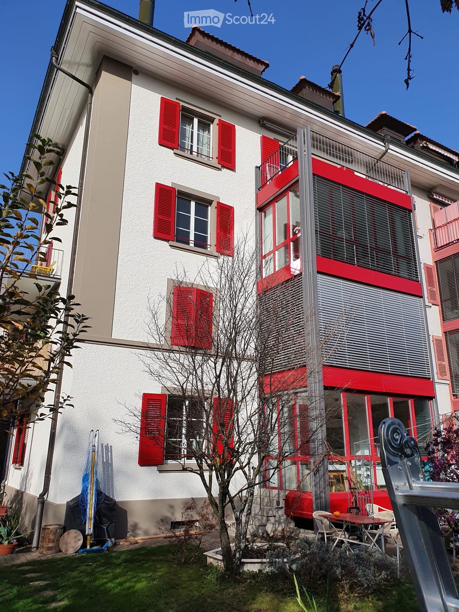 Two-story house with white walls, red shutters, and a balcony. The house features a small garden with a table, chairs, and a slide.