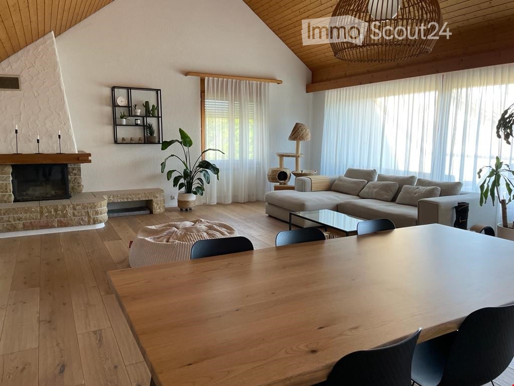 Modern living room with wooden floors, a stone fireplace, large windows with white curtains, a grey sectional sofa, a glass coffee table, a wooden dining table with black chairs, a round ottoman, a cat tree, a floor lamp, and decorative shelves with plant