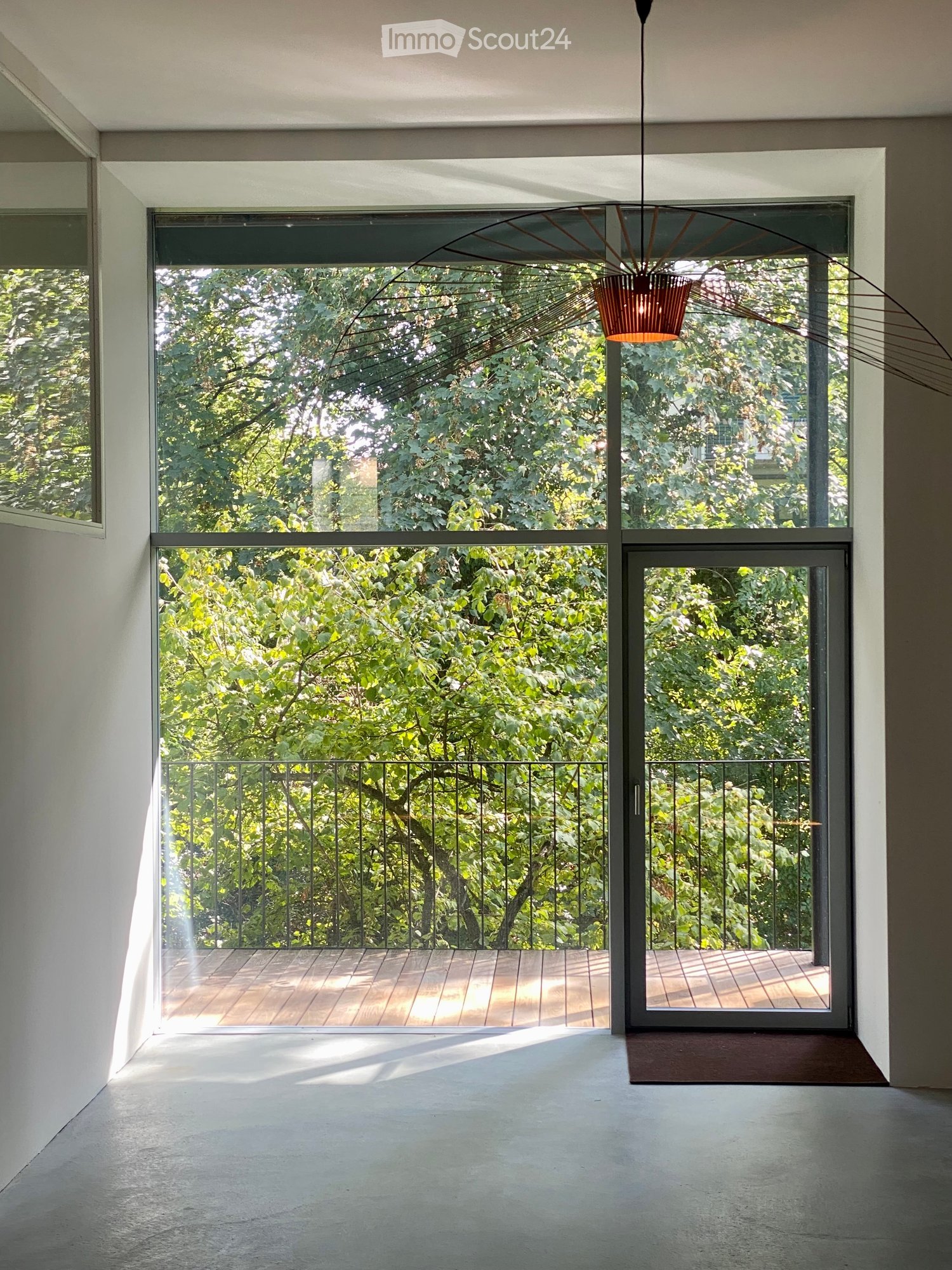 Modern balcony with wooden decking, black metal railing, large glass doors leading to the interior, and a view of lush greenery.