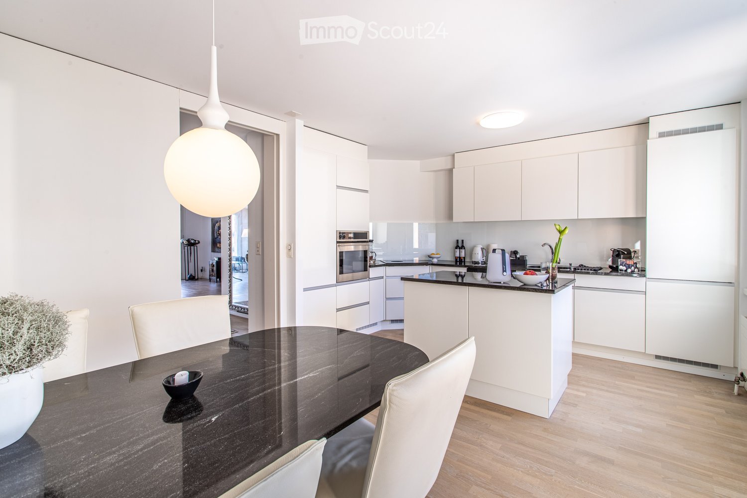 Modern kitchen with white cabinets, a central island, and a dining area featuring a dark wooden table and white chairs. The kitchen includes an oven, refrigerator, and various appliances on the counter.