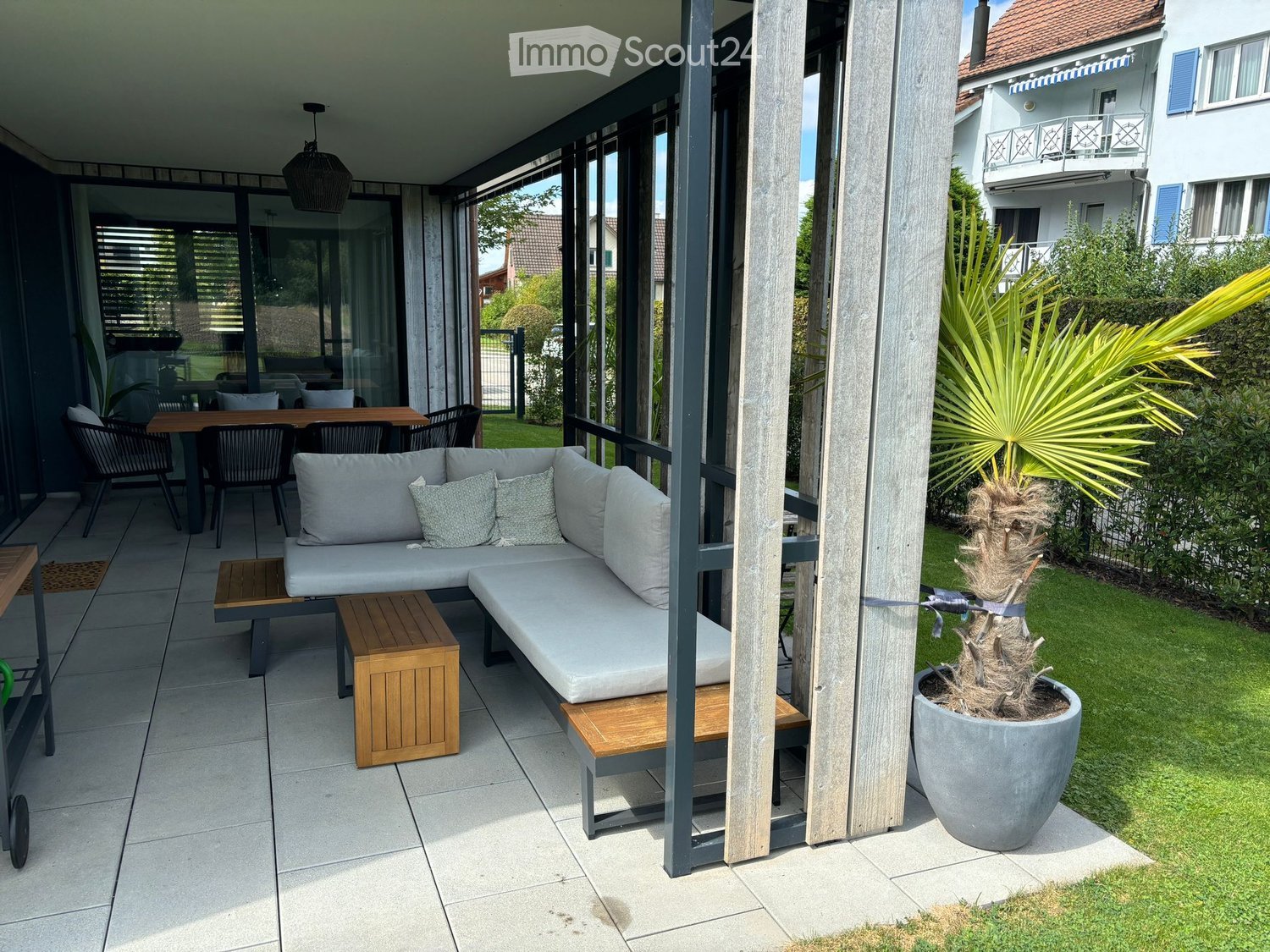 Balcony with a grey sectional sofa, wooden stools, a dining table, and chairs, surrounded by a wooden railing and a potted palm tree.