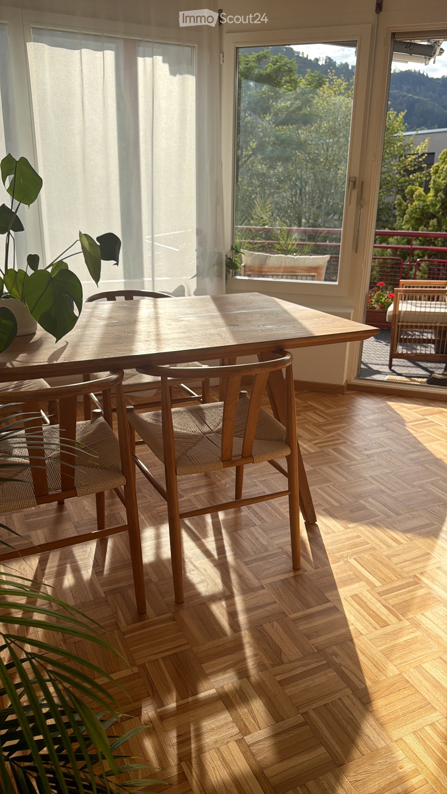 Bright dining area with wooden table, chairs, and a plant near the window, overlooking a scenic outdoor view.