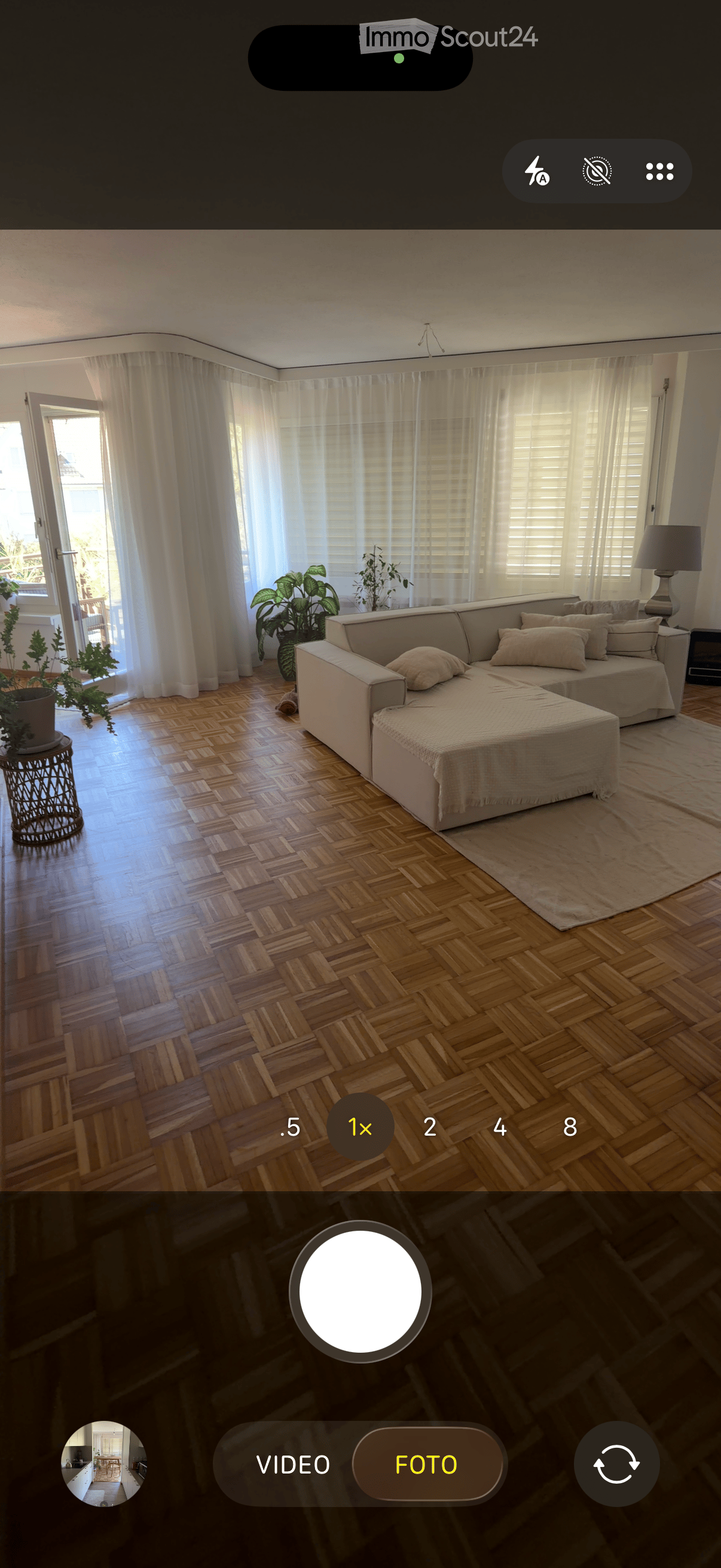 Bright living room with wooden herringbone flooring, large windows with white curtains, a cream-colored sectional sofa with multiple pillows, a side table with a table lamp, and potted plants near the windows.