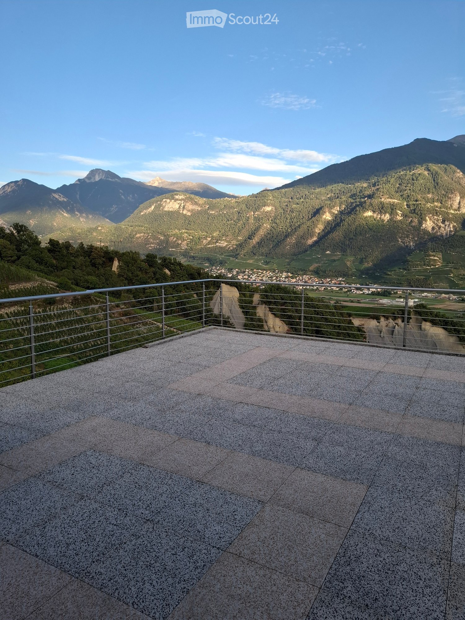 A spacious balcony with a modern metal railing, overlooking a scenic valley and mountains.