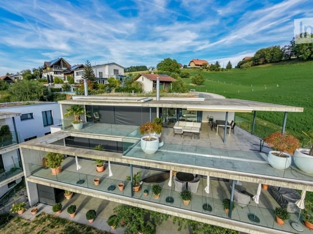 Modern two-story house with a large terrace and glass railings, surrounded by greenery and potted plants. The house features a flat roof and contemporary design.
