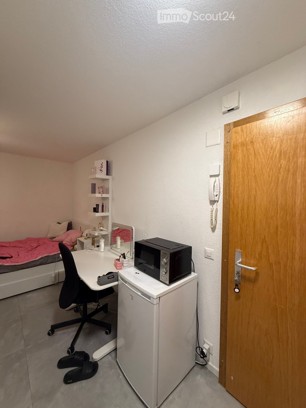 Small furnished room with a bed, desk, chair, microwave on a mini-fridge, shelf, mirror, and telephone on the wall.