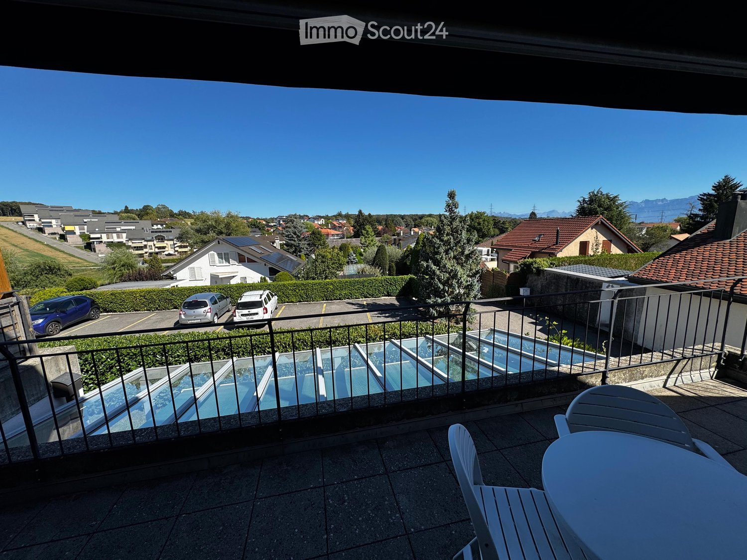 Balcony with a view of a residential area, featuring a table and chairs, a swimming pool, and a parking area with cars.