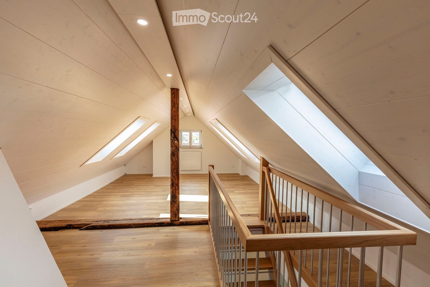 Bright, open attic space with wooden floors, exposed wooden beams, and skylights allowing natural light to flood the area. The space features a wooden staircase leading to an upper level, with a small window and radiator visible in the background.