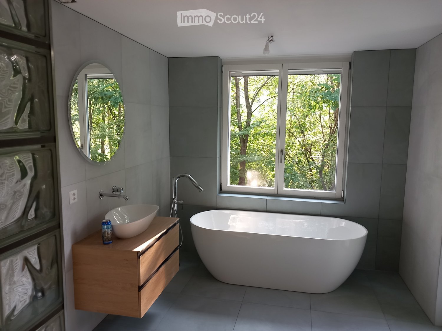 Modern bathroom with a large freestanding bathtub, a wooden sink cabinet, a modern faucet, and a round mirror. The room features large windows with a view of greenery outside.