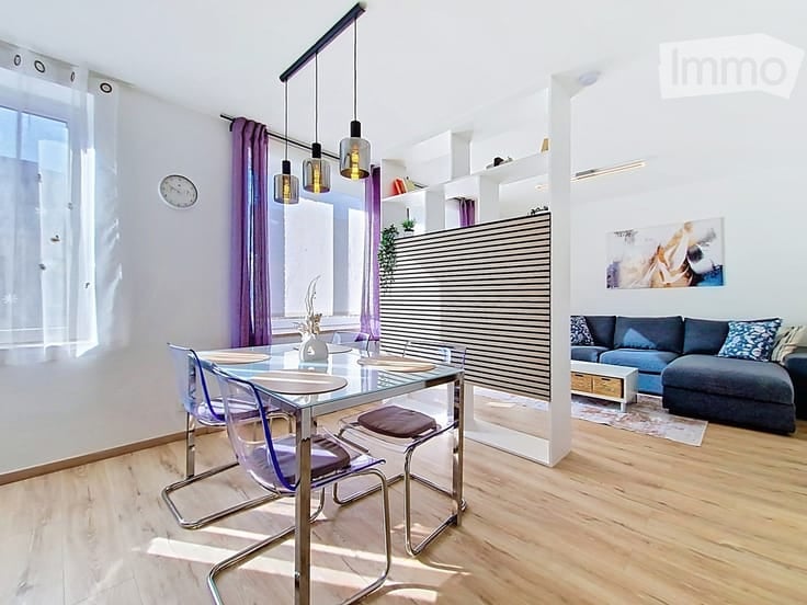 Modern open-plan living and dining area with light wood flooring, a blue sectional sofa, a glass dining table with four chairs, pendant lights, shelves, and artwork on the wall.