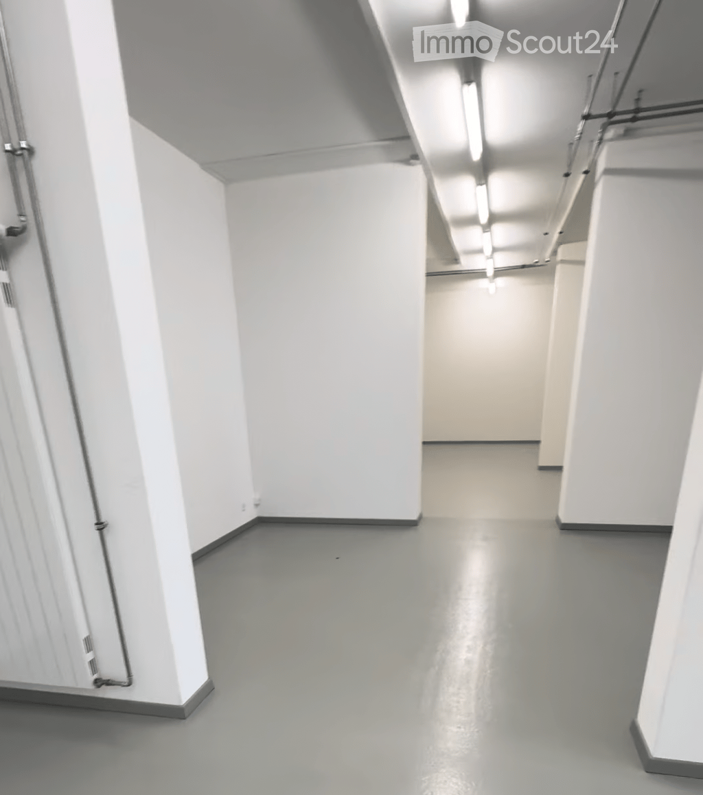 Empty corridor with white walls, grey floor, and industrial-style lighting. No furniture or equipment present.
