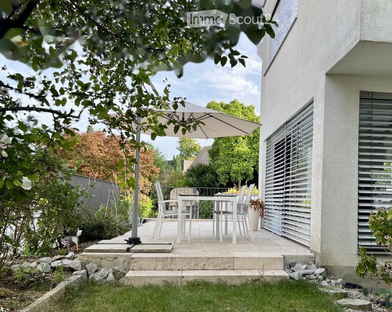 A modern outdoor patio area with a white dining set, umbrella, and surrounding greenery.