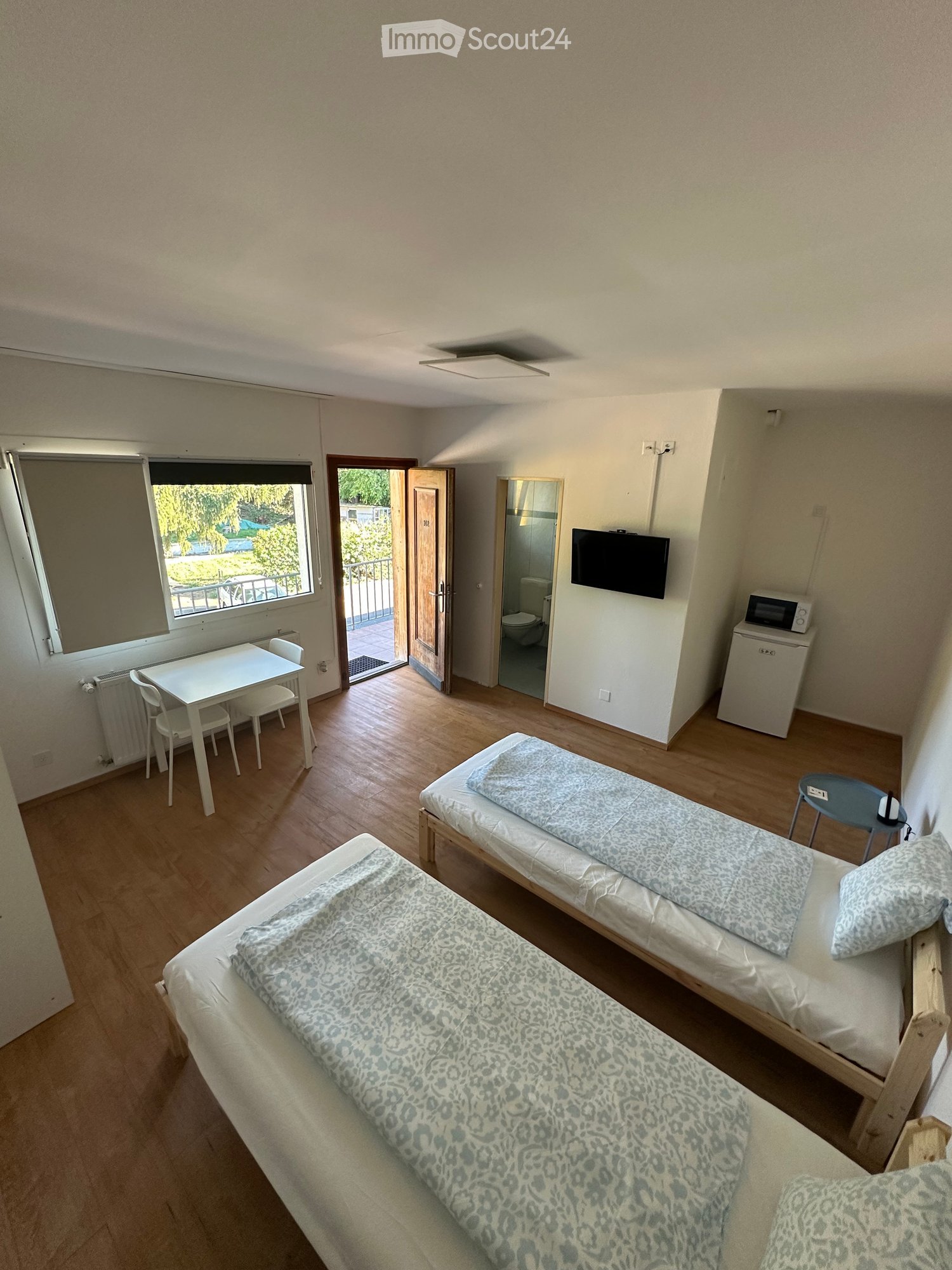 Cozy furnished room with two single beds, a small table with chairs, a microwave, a refrigerator, and a TV. The room has wooden flooring and a window with a view of the outdoor area.