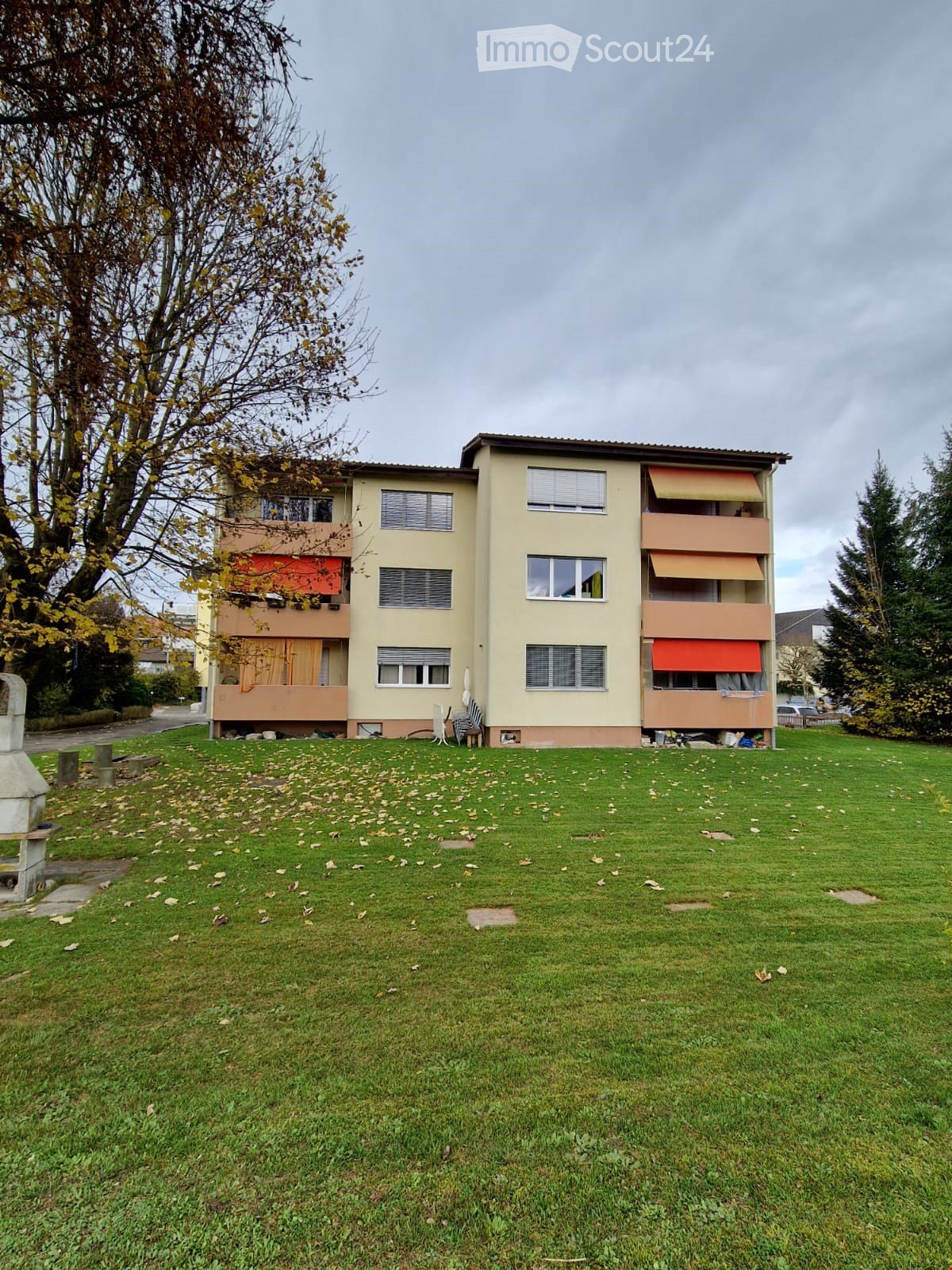 A three-story residential building with beige walls, multiple windows, and balconies with orange awnings. The building is surrounded by a grassy lawn with scattered leaves and trees.