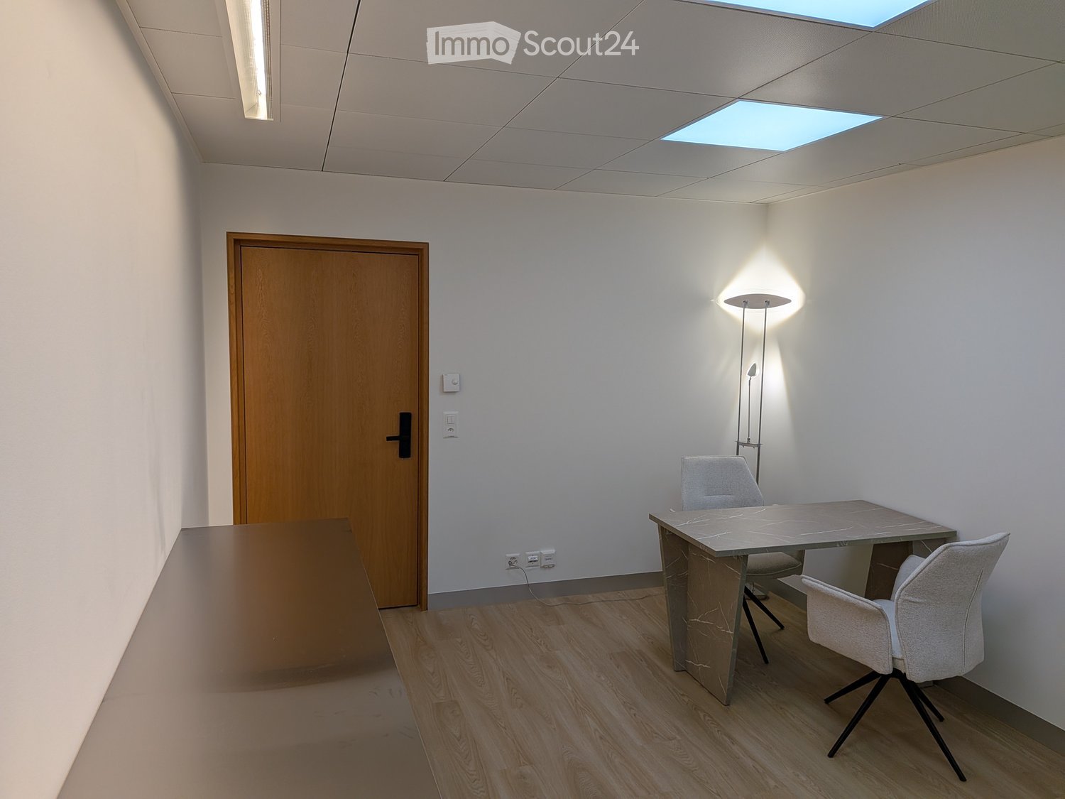 A modern, minimalist office space with a wooden floor, white walls, and a skylight. It features a sleek table with two chairs and a floor lamp.
