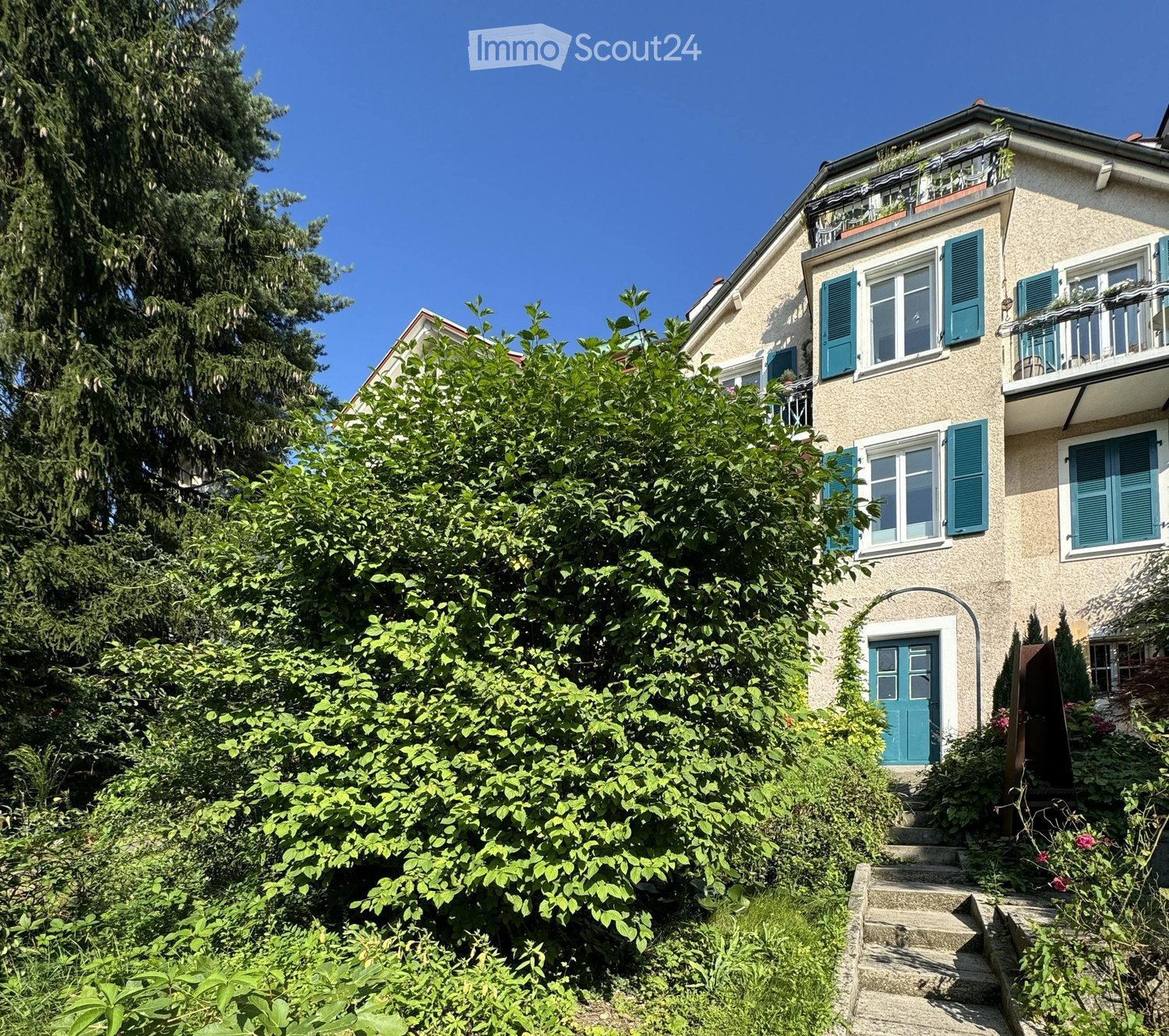 Two-story house with beige siding, green shutters, and a balcony on the upper level. The house is surrounded by lush greenery and trees.