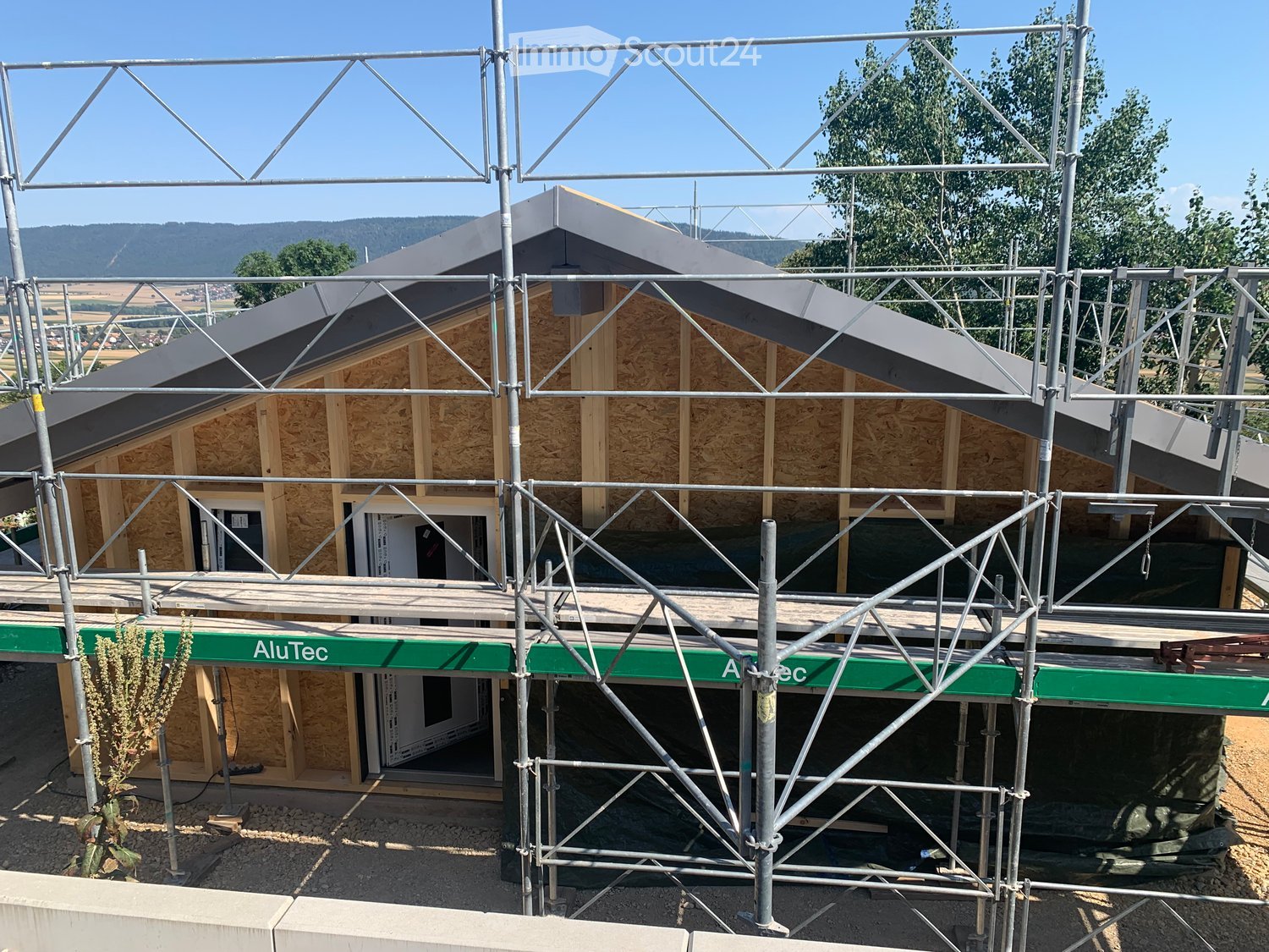 A partially constructed house with exposed wooden framing and metal scaffolding, featuring a grey roof and a visible window frame.