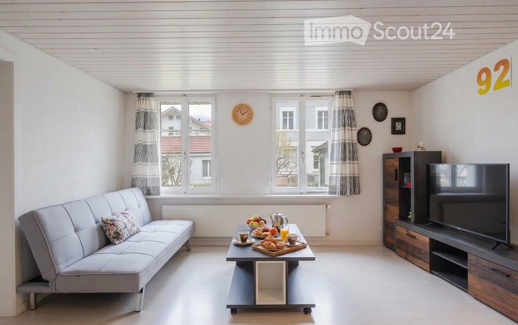 Bright living room with white walls, large windows, grey sofa, coffee table with breakfast items, television, and decorative wall clocks.