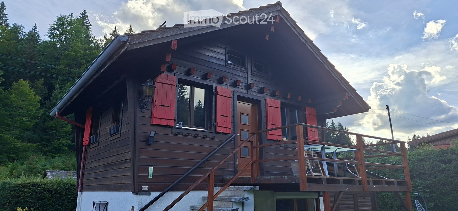 A two-story wooden house with red shutters, a balcony, and a staircase leading to the entrance. The house is surrounded by trees and has a rustic, alpine-style design.