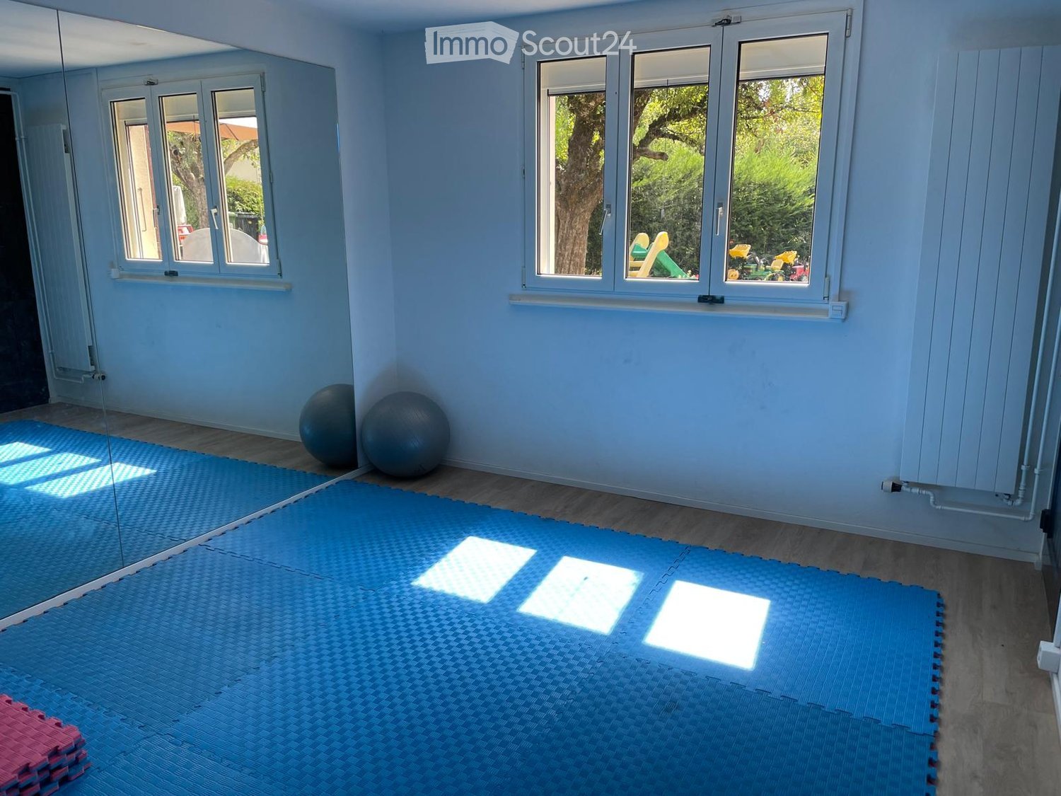 Bright room with light blue walls, wooden floor, large windows, exercise mat, fitness balls, mirror, and foam blocks.