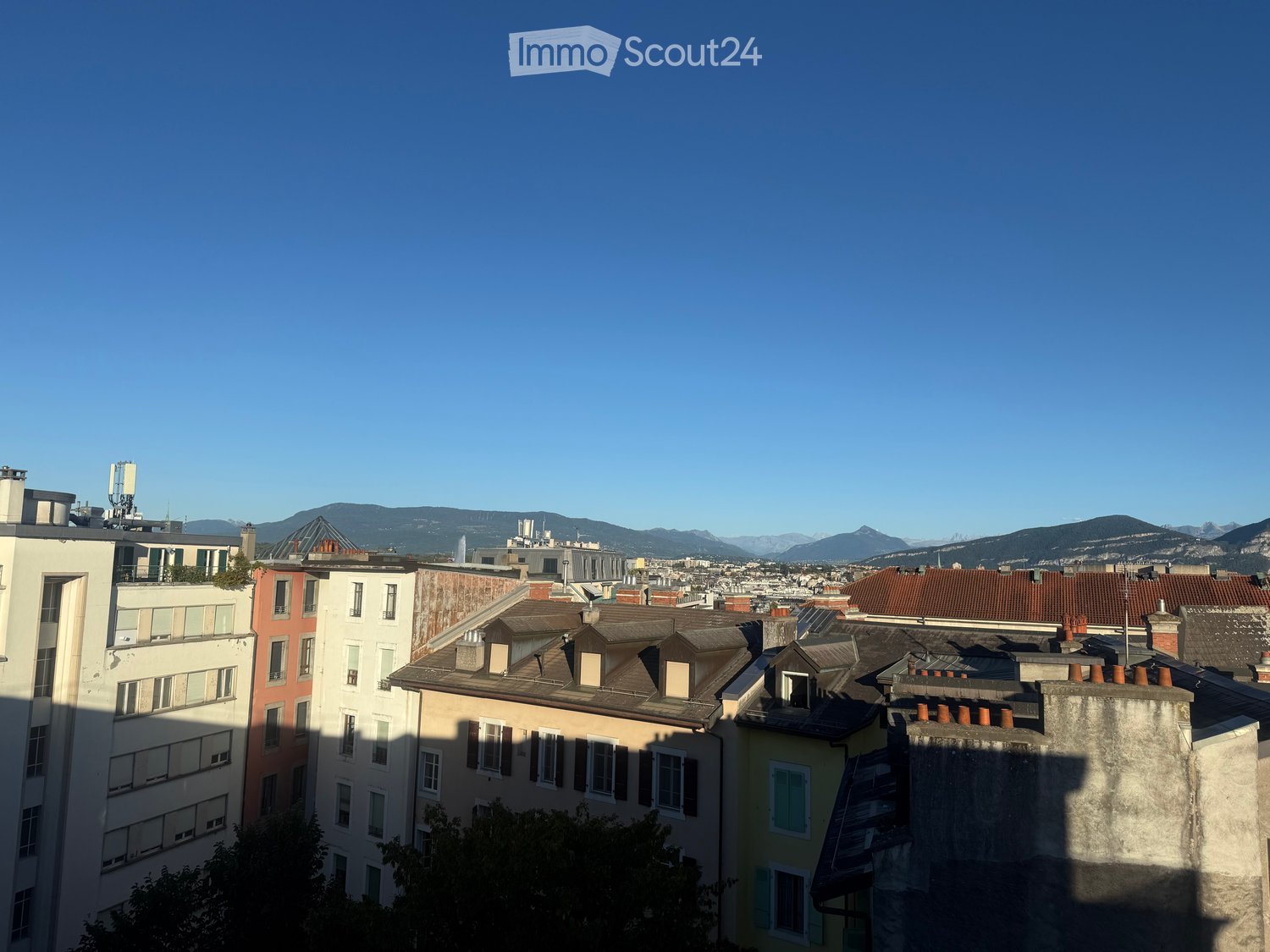 A cityscape view showing multiple buildings with varied roof styles and colors, set against a backdrop of distant mountains and a clear blue sky.