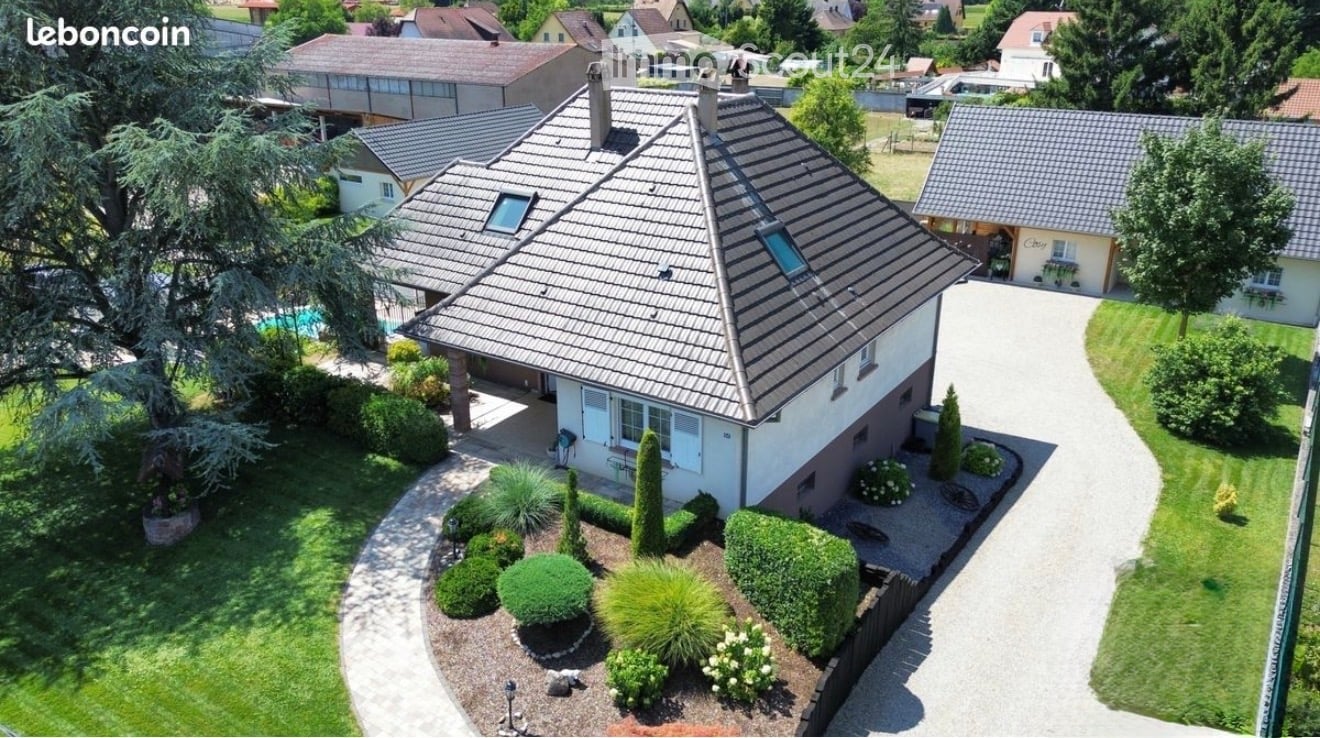 Aerial view of a two-story house with a gray tiled roof, surrounded by a well-maintained garden with shrubs and trees. A swimming pool is visible in the background, along with neighboring houses and a driveway leading to the property.