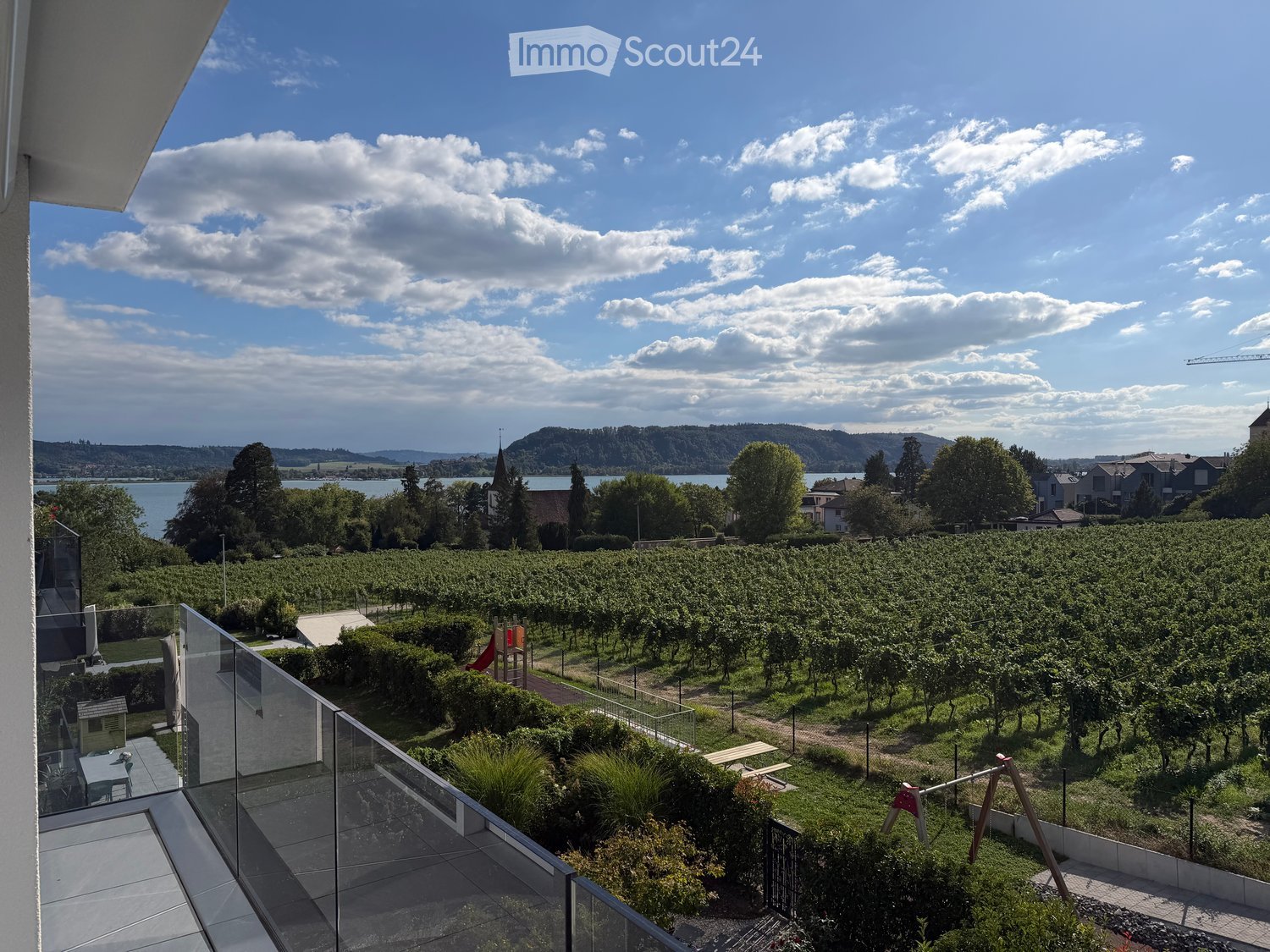 Balcony overlooking a vineyard, playground, and scenic lake view with distant hills.