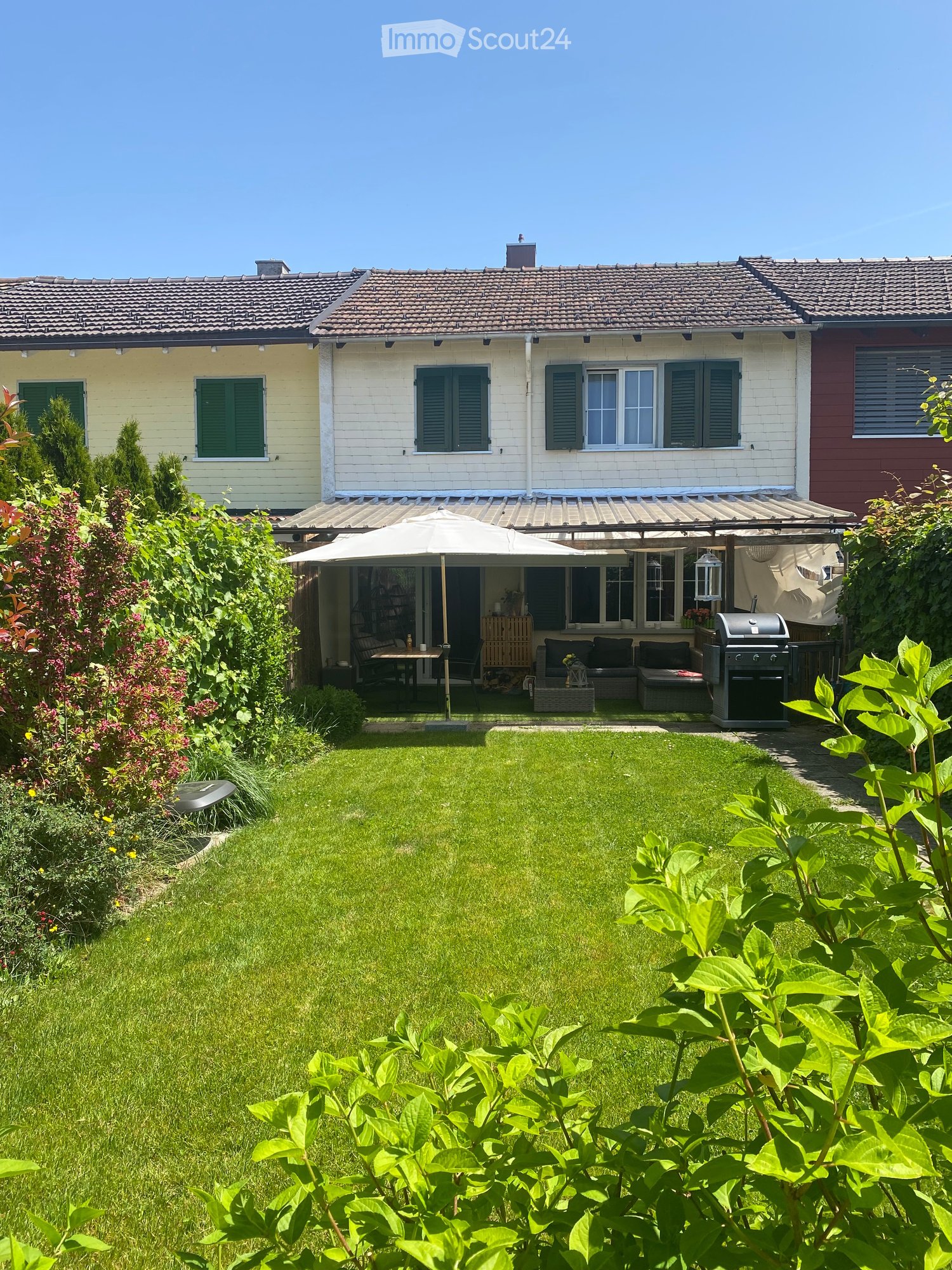 Well-maintained garden with a lawn, patio seating area, umbrella, grill, and surrounding greenery.