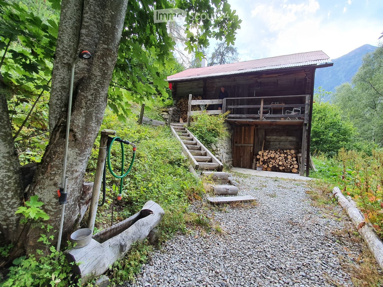 A wooden cabin with a red roof and balcony, surrounded by greenery and mountains. Stairs lead up to the cabin, and there is a stack of firewood and a hose nearby.