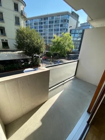 Balcony with a modern design, featuring a concrete floor and glass railing, overlooking a cityscape with trees and buildings.
