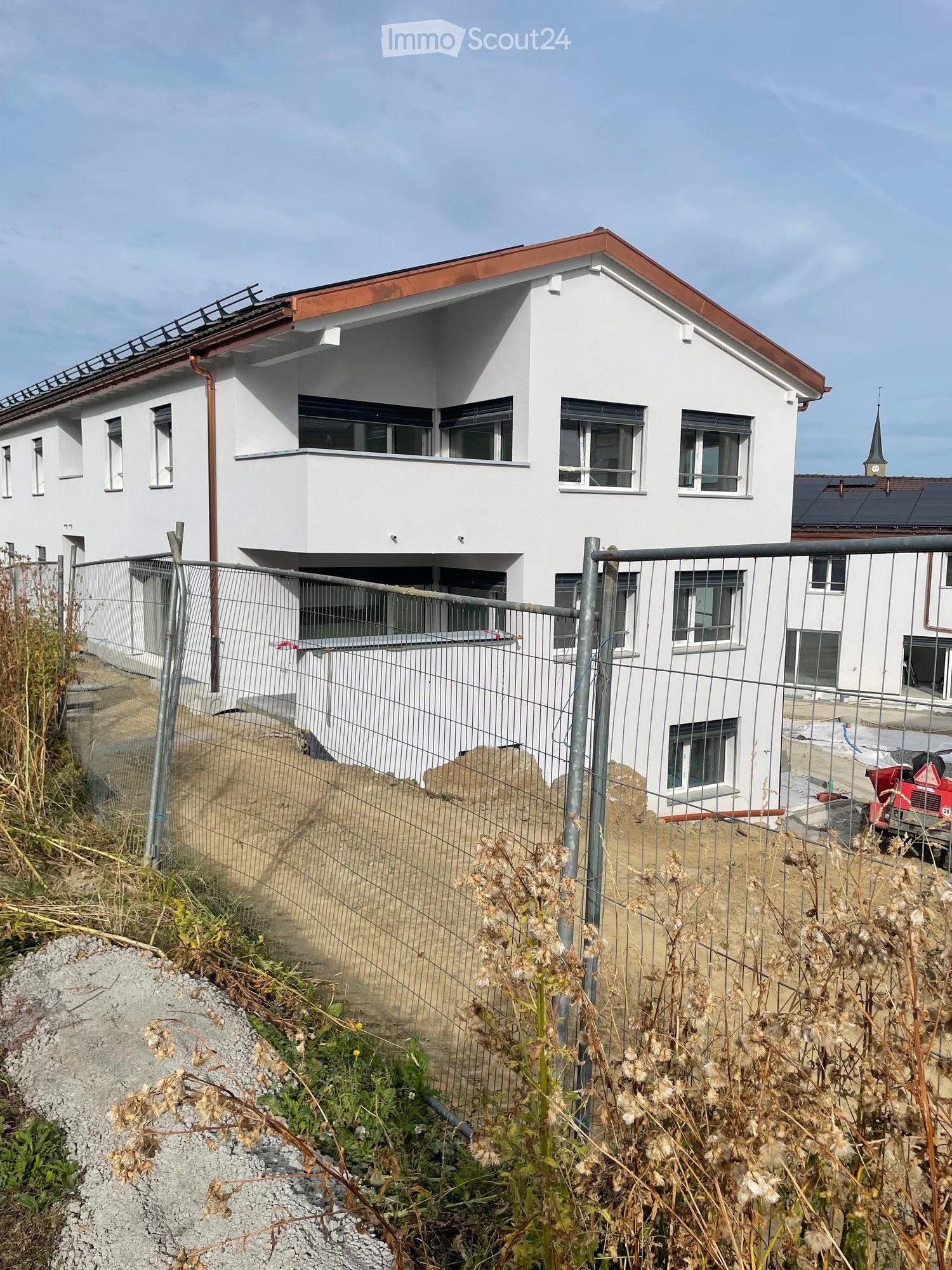 Modern two-story white house with a red roof, multiple windows, and a fenced yard with construction materials and equipment visible.