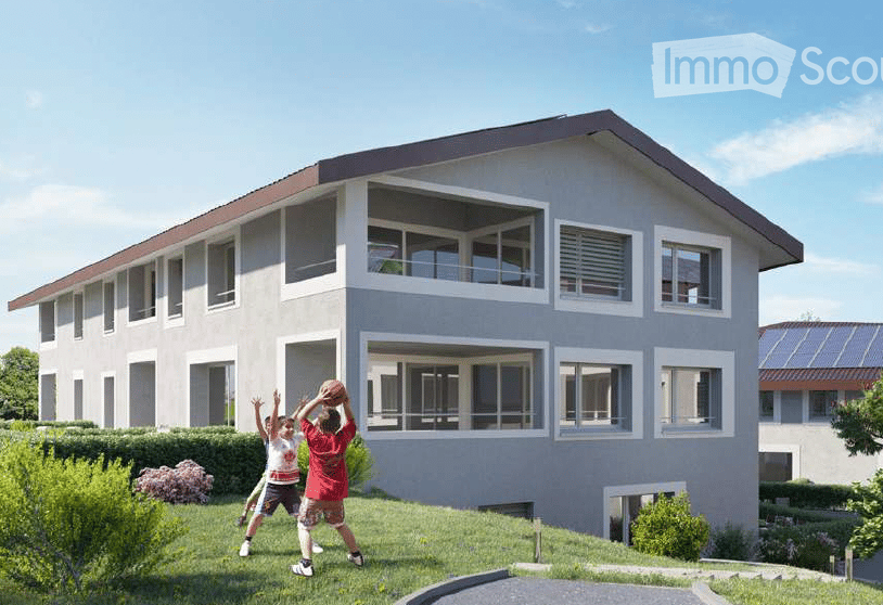 Modern two-story house with light gray exterior, large windows, and a balcony on the upper floor. The house features a sloped roof with dark roofing material and is surrounded by a well-maintained lawn and garden area.