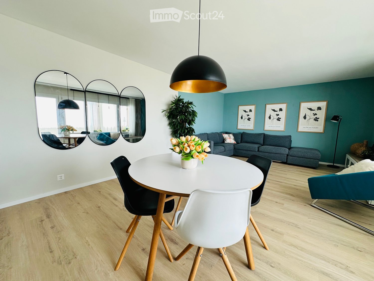 Modern living room with a round white dining table, black and white chairs, grey sofa, teal armchair, large pendant light, and decorative mirrors.