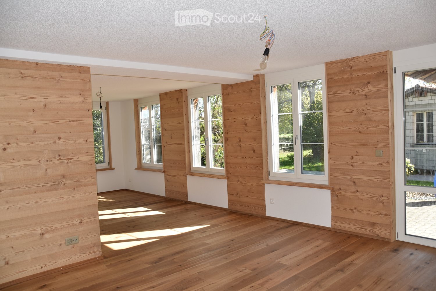 Empty room with wooden flooring and white walls, featuring multiple windows and a sliding glass door leading outside.