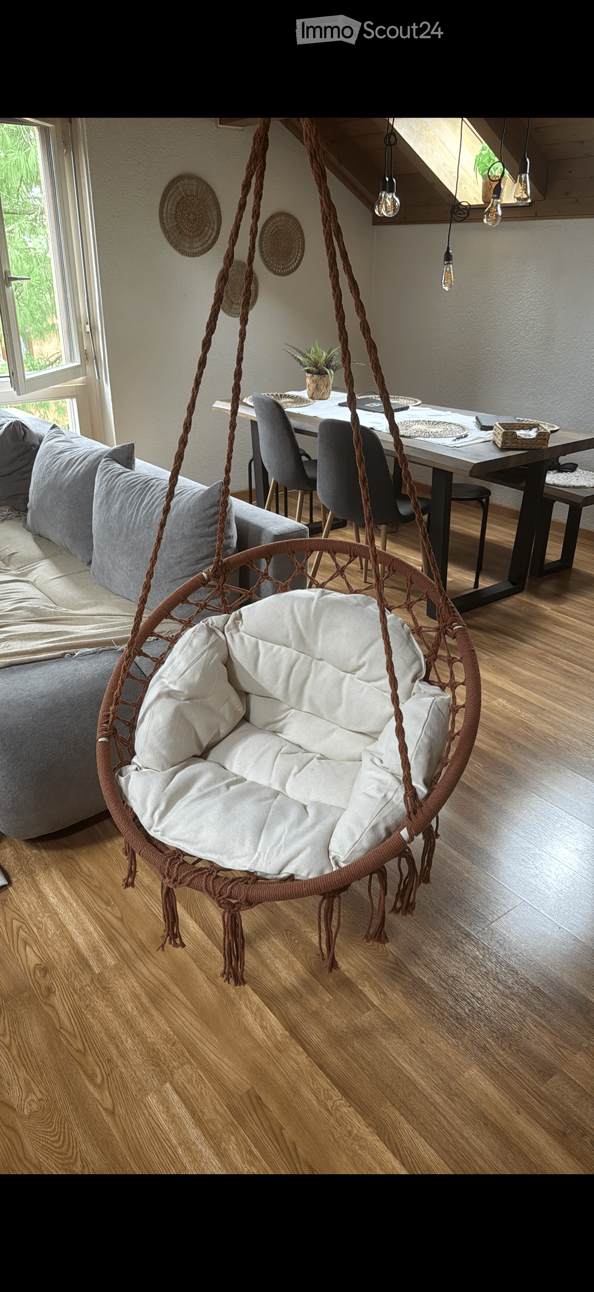 A cozy living space with a large grey sofa, a wicker hanging chair with white cushions, a modern dining table with chairs, and a small potted plant on the table. The room features wooden flooring and large windows with a view of greenery.