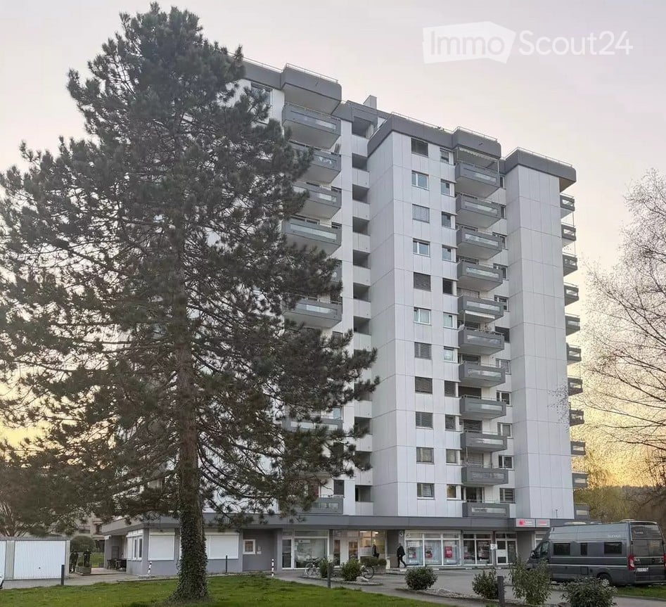 Modern multi-story apartment building with balconies, surrounded by trees and a small landscaped area.