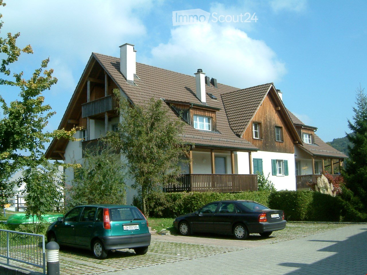 Two-story house with a brown tiled roof, white walls, and multiple windows. Features a balcony on the upper floor and a driveway with two parked cars.